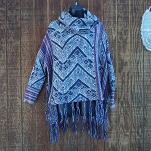 May include: A blue, white, and red patterned poncho with a cowl neck and fringe. The poncho has a geometric design and is hanging on a black hanger against a wooden background.