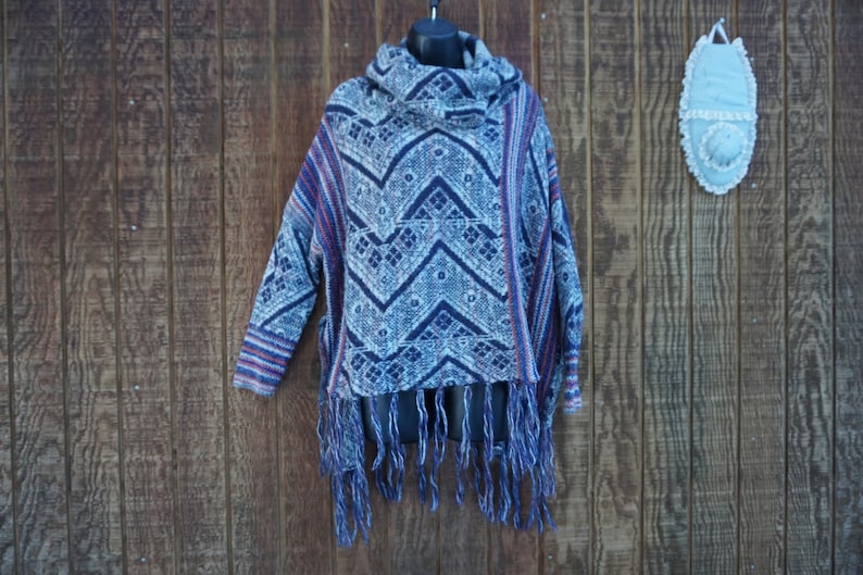 May include: A blue, white, and red patterned poncho with a cowl neck and fringe. The poncho has a geometric design and is hanging on a black hanger against a wooden background.