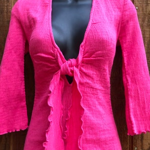 May include: A bright pink top with a plunging neckline and long, flared sleeves. The top features a tie detail at the waist and ruffled edges along the front. The fabric has a textured appearance.