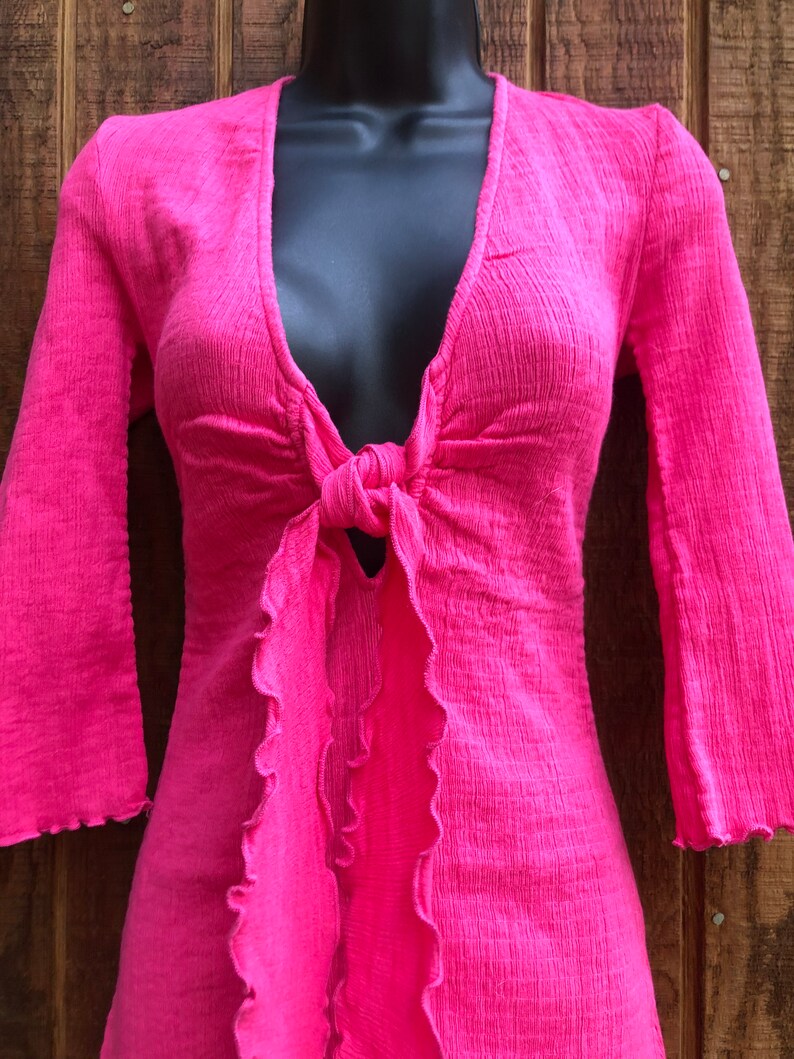May include: A bright pink top with a plunging neckline and long, flared sleeves. The top features a tie detail at the waist and ruffled edges along the front. The fabric has a textured appearance.