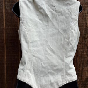 Puede incluir: Un chaleco sin mangas beige claro con cuello marr&oacute;n oscuro. El chaleco se muestra en una percha negra. La parte trasera del chaleco es visible, mostrando costuras a medida y un dobladillo curvo. Fondo de madera.