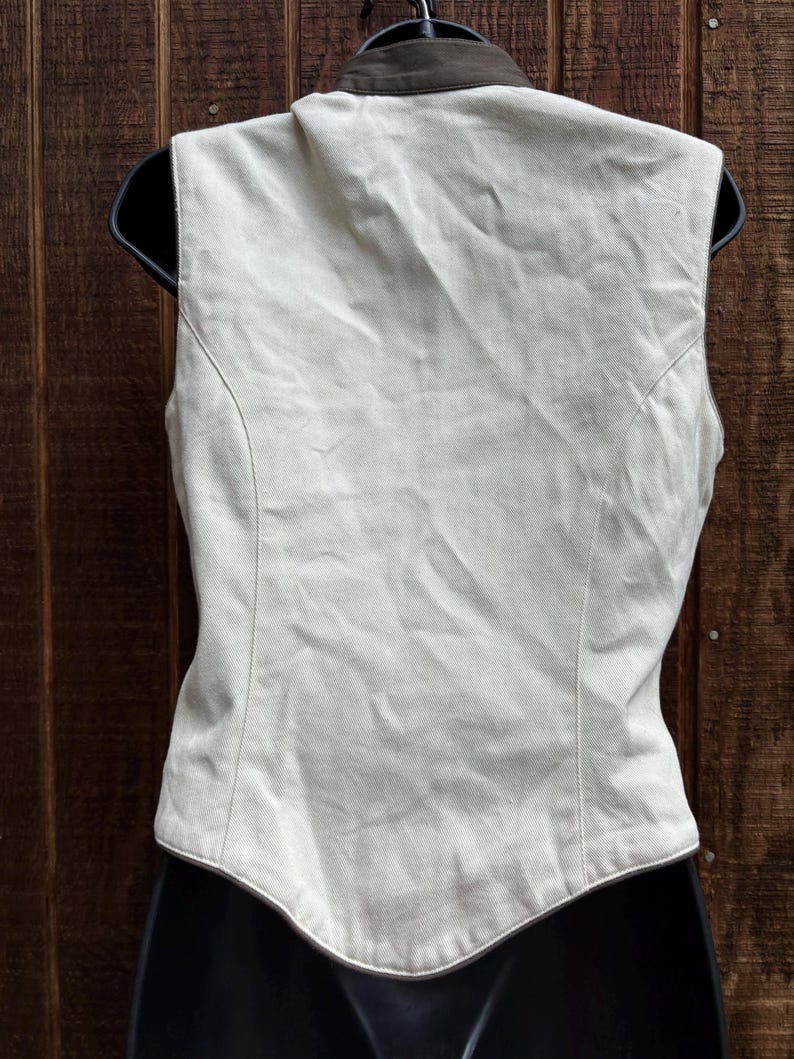 Puede incluir: Un chaleco sin mangas beige claro con cuello marr&oacute;n oscuro. El chaleco se muestra en una percha negra. La parte trasera del chaleco es visible, mostrando costuras a medida y un dobladillo curvo. Fondo de madera.