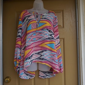 May include: A long-sleeved tunic top with a vibrant, colorful pattern. The top features a pink, yellow, blue, and white geometric design. It has a V-neckline with a tie closure and is displayed on a mannequin.