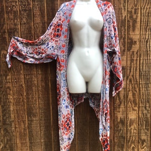 May include: A sheer, open-front kimono-style cover-up with a floral pattern in red, blue, and orange on a white background. The garment is displayed on a white mannequin against a wooden backdrop. The sleeves are long and flowing.