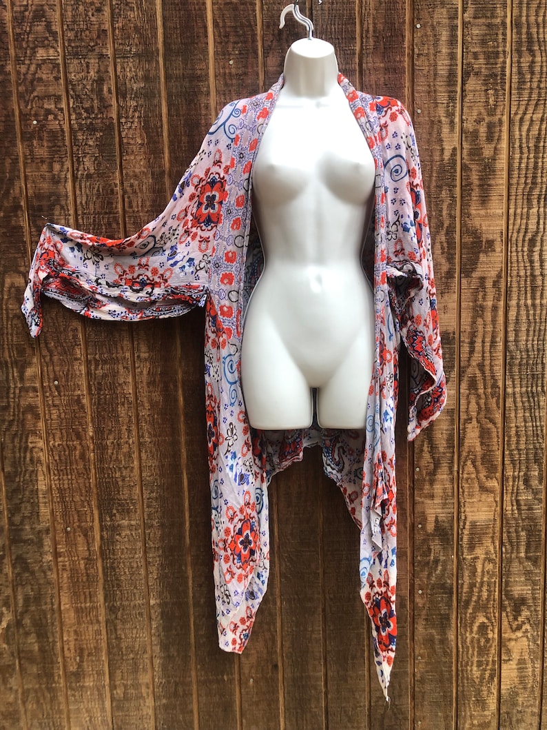May include: A sheer, open-front kimono-style cover-up with a floral pattern in red, blue, and orange on a white background. The garment is displayed on a white mannequin against a wooden backdrop. The sleeves are long and flowing.