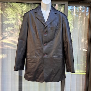 May include: A black leather blazer with a classic three-button closure and notched lapels. The jacket features a breast pocket and two side pockets. The blazer is displayed on a mannequin against a backdrop of a glass door and wooden paneling.