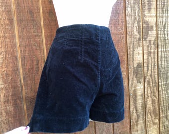 Puede incluir: Pantalones cortos de cintura alta azul marino hechos de un material de terciopelo suave. Los pantalones cortos tienen un ajuste a medida con una cintura alta y una cintura estructurada. Los pantalones cortos se muestran sobre un fondo de madera.