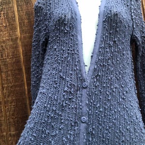 K&ouml;nnte beinhalten: Ein periwinkelfarbener blauer Cardigan mit einem strukturierten, noppenartigen Strickdesign. Der lang&auml;rmlige Pullover hat einen V-Ausschnitt und drei Kn&ouml;pfe auf der Vorderseite. Der Cardigan wird auf einer Schaufensterpuppe vor einem Holzgrund gezeigt.