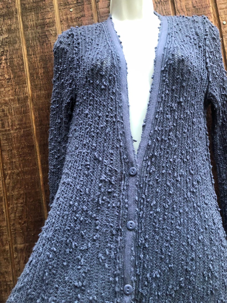 K&ouml;nnte beinhalten: Ein periwinkelfarbener blauer Cardigan mit einem strukturierten, noppenartigen Strickdesign. Der lang&auml;rmlige Pullover hat einen V-Ausschnitt und drei Kn&ouml;pfe auf der Vorderseite. Der Cardigan wird auf einer Schaufensterpuppe vor einem Holzgrund gezeigt.
