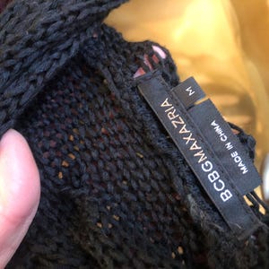 以下が含まれることがあります： 黒色のニット衣料品のクローズアップ。織り目が特徴的です。衣料品には「BCBGMAXAZRIA」と「MADE IN CHINA」と書かれたタグが付いています。サイズは「M」と表示されています。ニットウェアはセーターまたはトップスのように見えます。