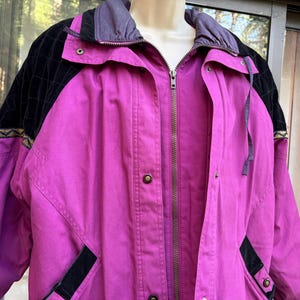 May include: A magenta jacket with a black quilted shoulder detail and a gray hood. The jacket has a zipper closure, snap buttons, and black accents on the pockets. The jacket has a geometric pattern on the shoulder.