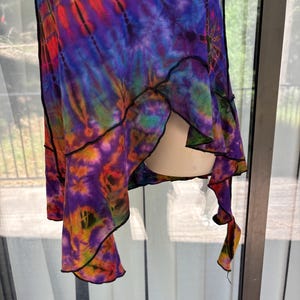 May include: A sleeveless garment featuring a vibrant tie-dye pattern in shades of purple, blue, orange, and red. The piece has an asymmetrical hemline with exposed black stitching along the edges, creating a layered, flowing silhouette.