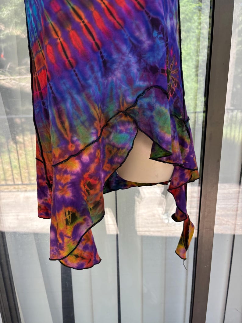 May include: A sleeveless garment featuring a vibrant tie-dye pattern in shades of purple, blue, orange, and red. The piece has an asymmetrical hemline with exposed black stitching along the edges, creating a layered, flowing silhouette.