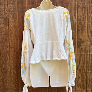 May include: White blouse with long sleeves and a peplum waist. The sleeves are adorned with yellow and blue floral embroidery. The blouse has a round neckline with a light blue trim and tie details at the cuffs.
