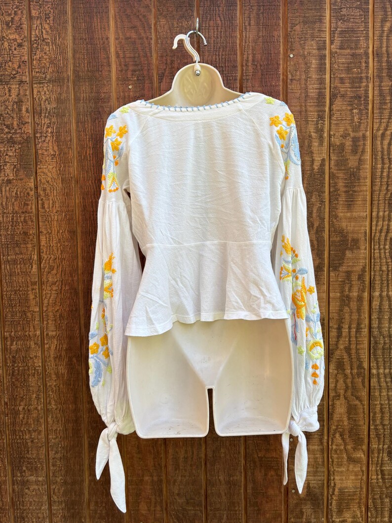 May include: White blouse with long sleeves and a peplum waist. The sleeves are adorned with yellow and blue floral embroidery. The blouse has a round neckline with a light blue trim and tie details at the cuffs.