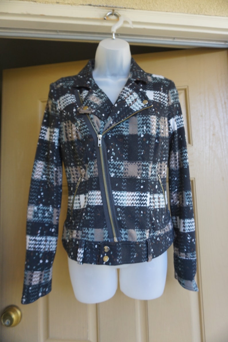 May include: A patterned jacket with a black, white, and blue plaid design. The jacket features a diagonal zipper, two front pockets, and gold-colored buttons. The jacket is displayed on a white mannequin.