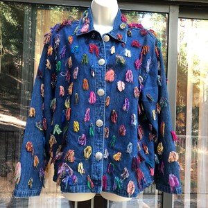 May include: A denim jacket with colorful, textured embellishments. The jacket is a medium blue wash with silver buttons and is adorned with various colored yarn accents, creating a unique, artistic look. The jacket has a classic collar and button-down front.