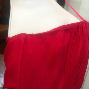 K&ouml;nnte beinhalten: Ein leuchtend rotes tr&auml;gerloses Kleid mit Perlenbesatz an der Taille. Das Kleid hat ein flie&szlig;endes Design und einen zarten Spaghettitr&auml;ger. Das Oberteil ist figurbetont, und der Rock scheint aus einem weichen, drapierenden Stoff zu sein.