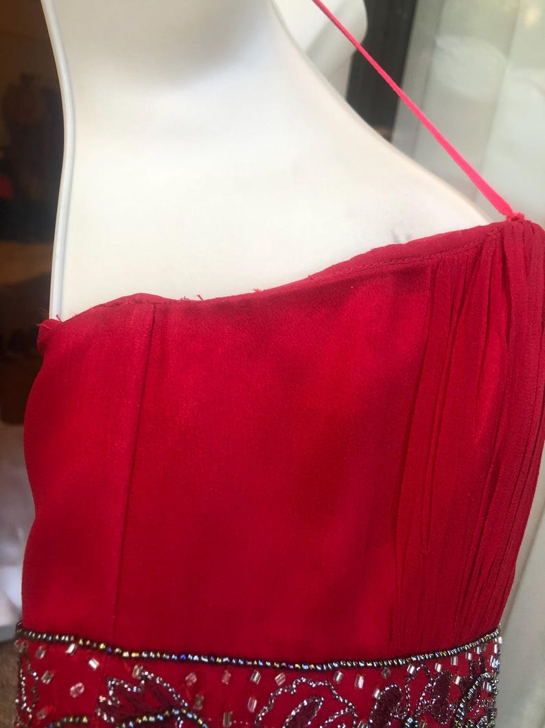 K&ouml;nnte beinhalten: Ein leuchtend rotes tr&auml;gerloses Kleid mit Perlenbesatz an der Taille. Das Kleid hat ein flie&szlig;endes Design und einen zarten Spaghettitr&auml;ger. Das Oberteil ist figurbetont, und der Rock scheint aus einem weichen, drapierenden Stoff zu sein.