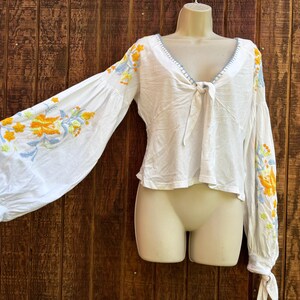 May include: White long-sleeve top with a V-neck and tie detail. The sleeves feature embroidered floral designs in yellow, blue, and orange. The top is displayed on a mannequin against a wooden background.