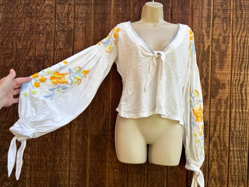 May include: White long-sleeve top with a V-neck and tie detail. The sleeves feature embroidered floral designs in yellow, blue, and orange. The top is displayed on a mannequin against a wooden background.