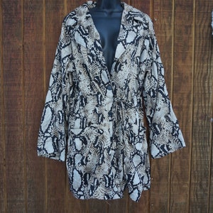 May include: A long-sleeved blazer with a snakeskin pattern in shades of black, beige, and white. The blazer has a tie waist, a notched collar, and two front pockets. The garment is displayed against a wooden background.