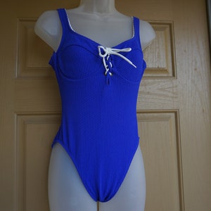 May include: A vibrant blue one-piece swimsuit with a textured pattern. The swimsuit features a lace-up detail at the bust and white trim along the straps and neckline. The swimsuit is displayed on a mannequin.