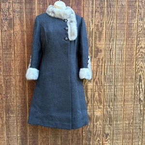 May include: A dark gray, vintage coat with a faux fur collar and cuffs. The coat has a button-up front and a flared skirt. The coat is hanging on a wooden hanger against a wooden background. The coat is a classic style.