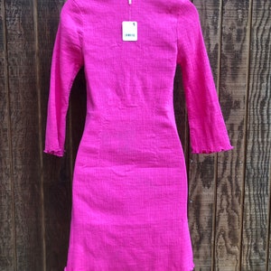 May include: A bright pink long-sleeved dress with a textured fabric. The dress has a fitted silhouette, a V-neck, and a scalloped hem and sleeve cuffs. A price tag hangs from the neckline.