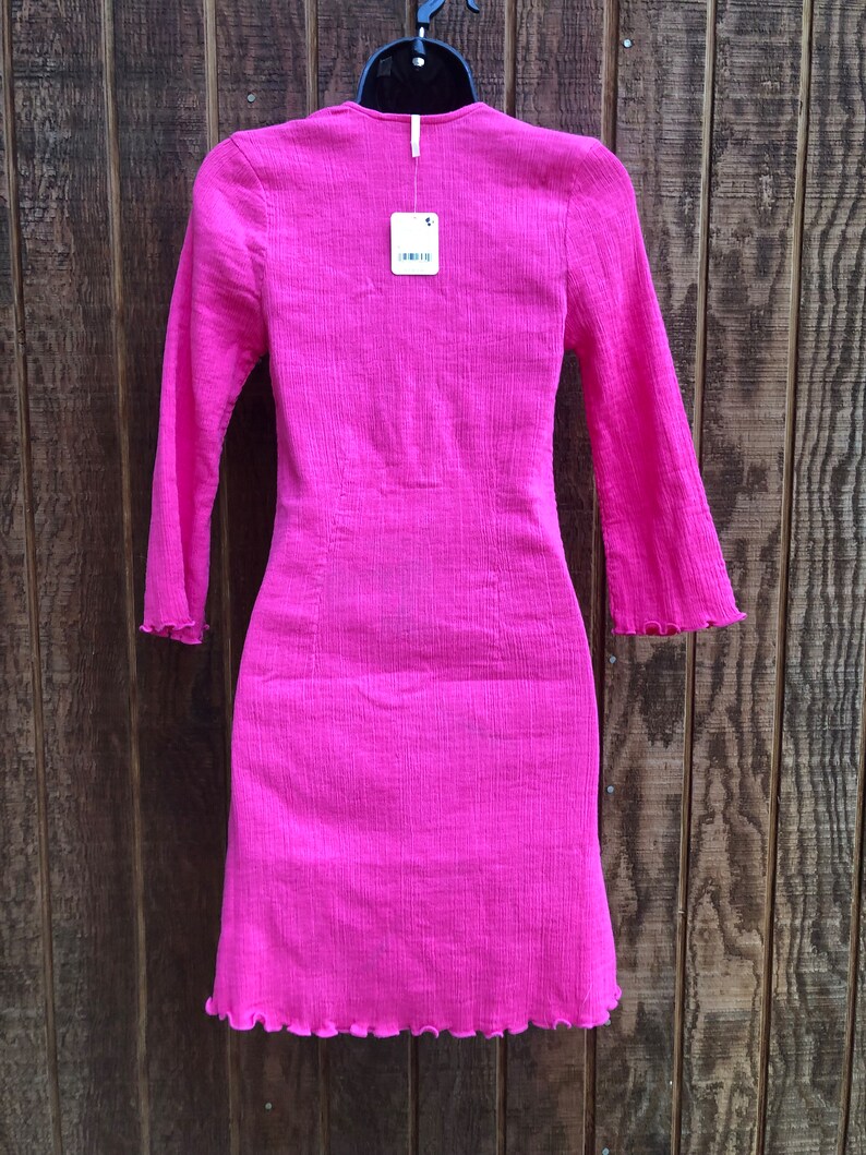 May include: A bright pink long-sleeved dress with a textured fabric. The dress has a fitted silhouette, a V-neck, and a scalloped hem and sleeve cuffs. A price tag hangs from the neckline.