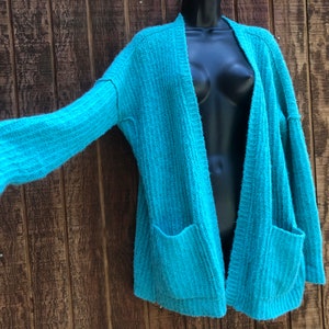 May include: A teal blue knitted cardigan with long sleeves and two pockets. The cardigan is open in the front and has a relaxed fit.