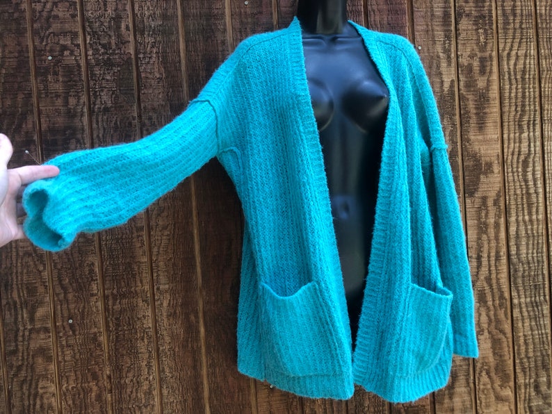 May include: A teal blue knitted cardigan with long sleeves and two pockets. The cardigan is open in the front and has a relaxed fit.