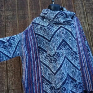 May include: A blue, white, and red patterned poncho-style sweater with a cowl neck. The sweater features a geometric design and striped accents on the sleeves and sides. The garment is displayed on a black hanger.