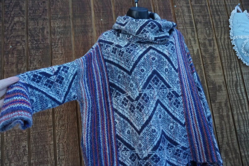 May include: A blue, white, and red patterned poncho-style sweater with a cowl neck. The sweater features a geometric design and striped accents on the sleeves and sides. The garment is displayed on a black hanger.