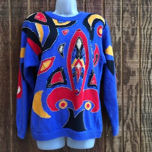 May include: A blue knitted sweater with a colorful abstract design featuring red, yellow, and black accents. The sweater is embellished with sequins and beads.