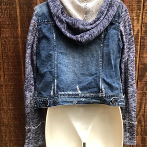 May include: A blue denim jacket with a gray knit hood and sleeves. The jacket has a distressed look with a button closure and is displayed on a white mannequin. The back of the jacket is visible, showcasing the design.