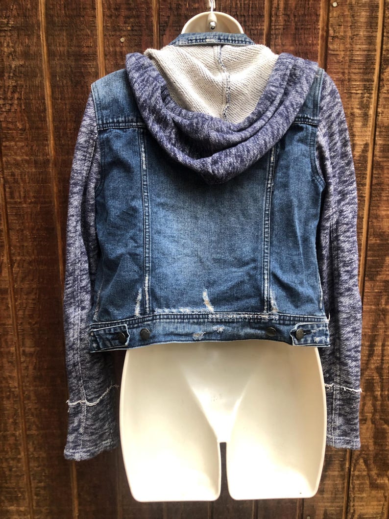 May include: A blue denim jacket with a gray knit hood and sleeves. The jacket has a distressed look with a button closure and is displayed on a white mannequin. The back of the jacket is visible, showcasing the design.