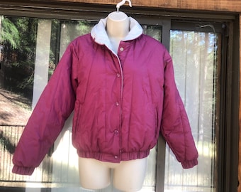 May include: A magenta quilted jacket with a white collar and white trim. The jacket has a button-up front and two pockets. The jacket is displayed on a mannequin. The background is a window with trees.