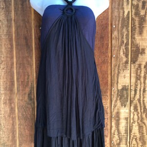 Op de afbeelding: Een marineblauwe halterjurk met een zwart ringdetail bij de halslijn. De jurk heeft een gelaagde rok en wordt getoond op een witte mannequin tegen een houten achtergrond. De jurk is mouwloos.