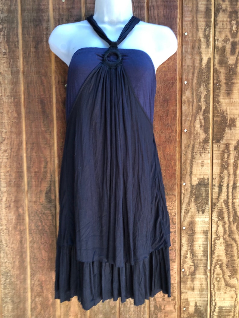 Op de afbeelding: Een marineblauwe halterjurk met een zwart ringdetail bij de halslijn. De jurk heeft een gelaagde rok en wordt getoond op een witte mannequin tegen een houten achtergrond. De jurk is mouwloos.