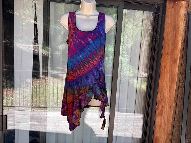 May include: A sleeveless tank top featuring a vibrant tie-dye pattern in shades of purple, blue, red, and orange. The garment has a scoop neckline and an asymmetrical hemline with a black stitched edge, displayed on a white mannequin torso.