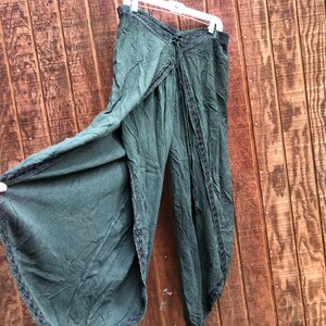 May include: Dark green palazzo pants with a unique wrap design. The pants are made of a soft, flowing fabric and feature decorative trim along the edges. They are hanging on a white hanger against a wooden background.