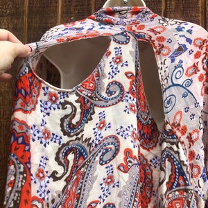 May include: A cream-colored, paisley-patterned top with orange, blue, and gray accents. The top has a unique open-back design and is displayed on a white mannequin. The fabric appears lightweight and flowing, suitable for warm weather.