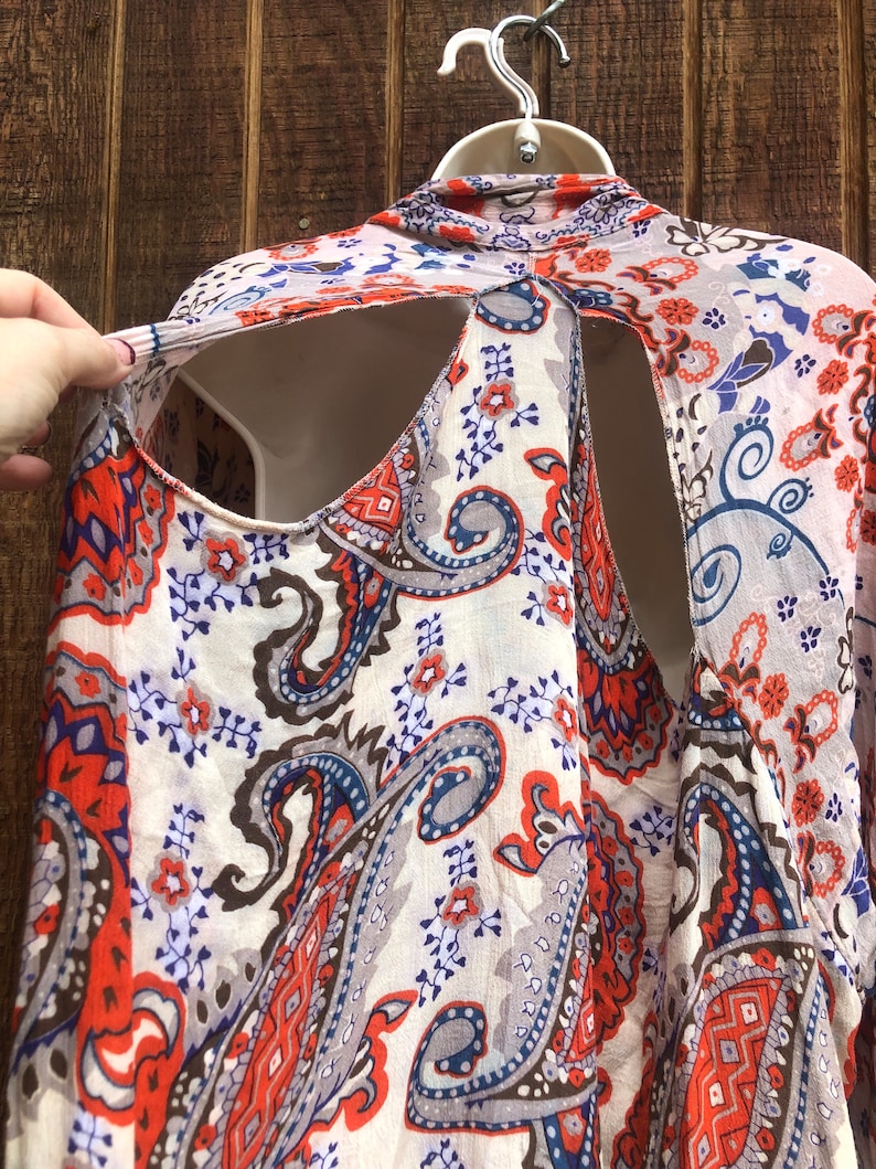 May include: A cream-colored, paisley-patterned top with orange, blue, and gray accents. The top has a unique open-back design and is displayed on a white mannequin. The fabric appears lightweight and flowing, suitable for warm weather.
