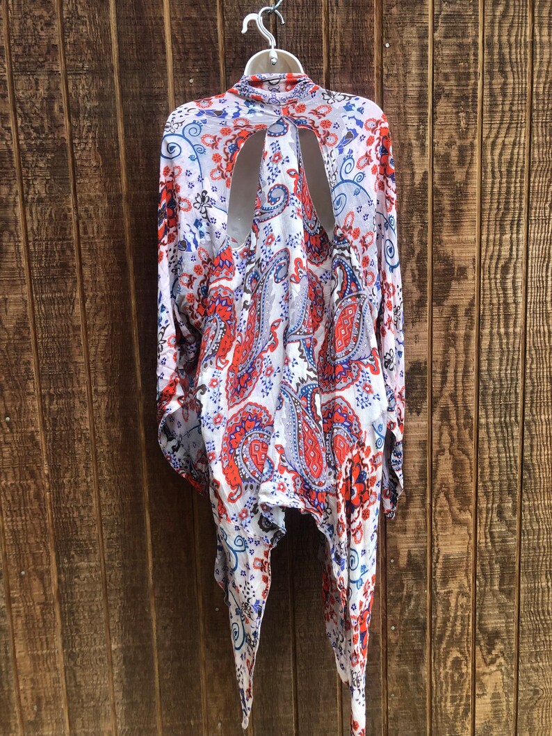 May include: A white, red, and blue paisley print kimono-style top with long sleeves and a unique open back design. The lightweight fabric hangs from a hanger against a wooden background.