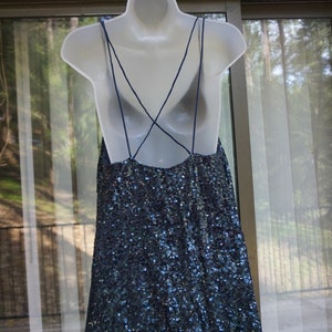 K&ouml;nnte beinhalten: Ein marineblaues Paillettenkleid mit Spaghettitr&auml;gern und tiefem R&uuml;ckenausschnitt wird auf einer wei&szlig;en Schaufensterpuppe pr&auml;sentiert. Das Kleid h&auml;ngt an einem wei&szlig;en Kleiderb&uuml;gel vor einem Fenster und B&auml;umen.