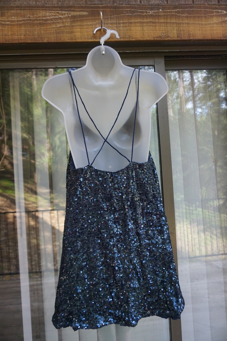 K&ouml;nnte beinhalten: Ein marineblaues Paillettenkleid mit Spaghettitr&auml;gern und tiefem R&uuml;ckenausschnitt wird auf einer wei&szlig;en Schaufensterpuppe pr&auml;sentiert. Das Kleid h&auml;ngt an einem wei&szlig;en Kleiderb&uuml;gel vor einem Fenster und B&auml;umen.