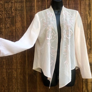 May include: A light pink, open-front jacket with long sleeves. The jacket features delicate floral embroidery in a matching pink thread. The sheer fabric drapes gracefully, creating a soft and elegant look. The jacket is displayed on a black mannequin.