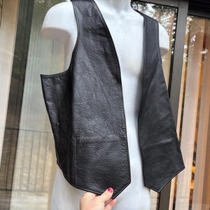 May include: A black leather vest with a textured grain finish, featuring a deep V-neckline, wide shoulder panels, and a pointed hemline. The garment is sleeveless and displays a clean, structured design with visible stitching along the edges.