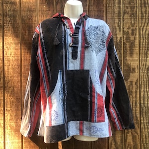 May include: A hooded pullover shirt with a unique patchwork design. The shirt features a mix of black, red, and blue striped patterns, along with a large front pocket and a decorative design on the front. The shirt is displayed on a mannequin against a wooden background.